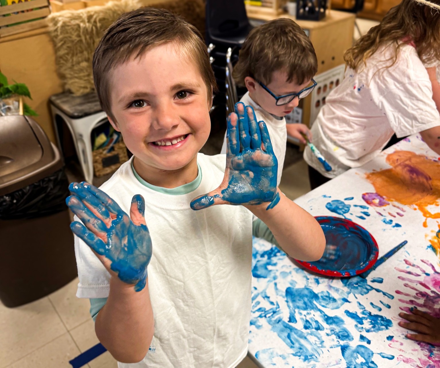 A young boy with blue paint on his hands, smiling, stands in a classroom. Behind him, a table displays more colorful paint.