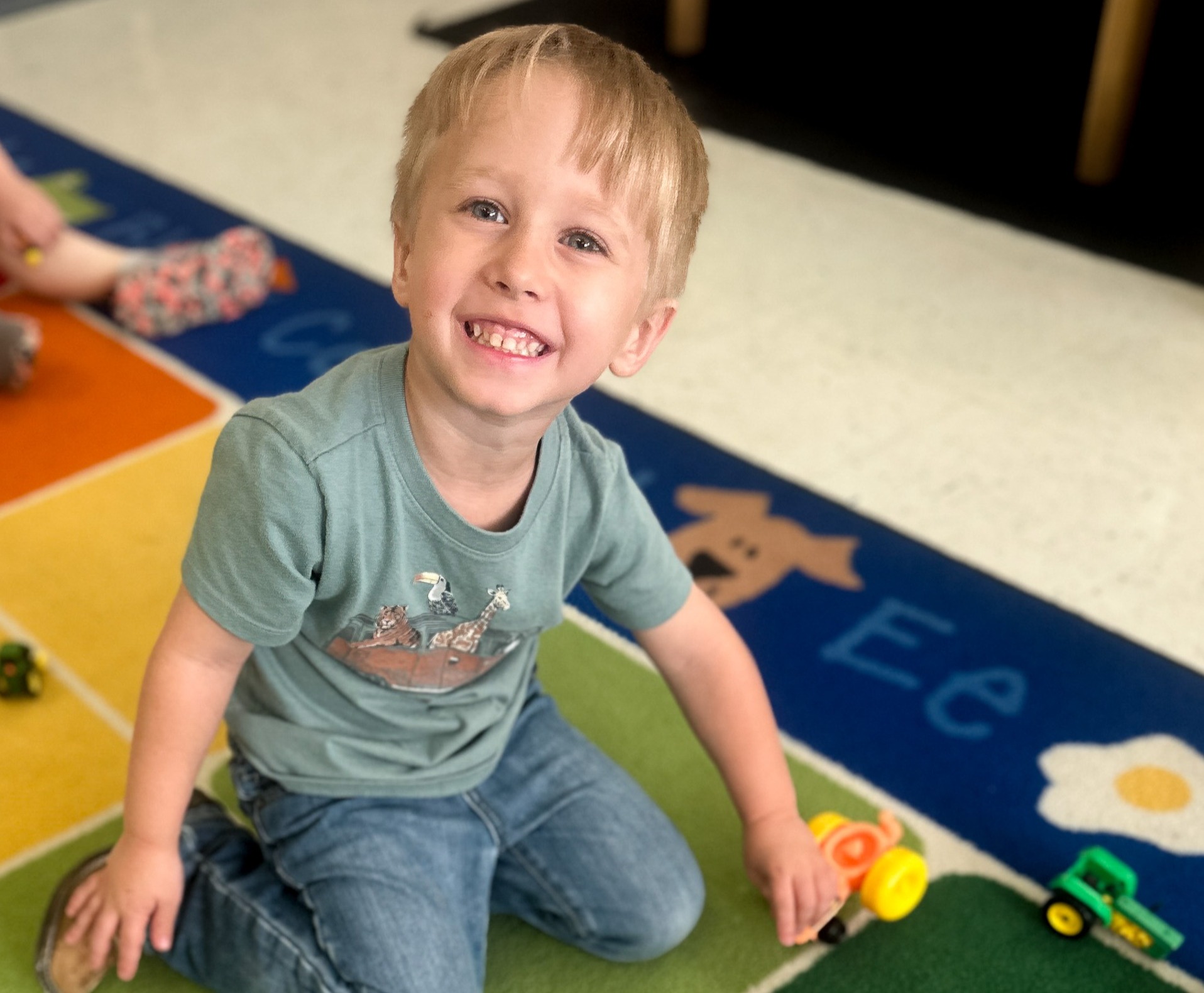 A young child smiles while playing with toys on a colorful carpet. Behind them is a rug with alphabet letters.