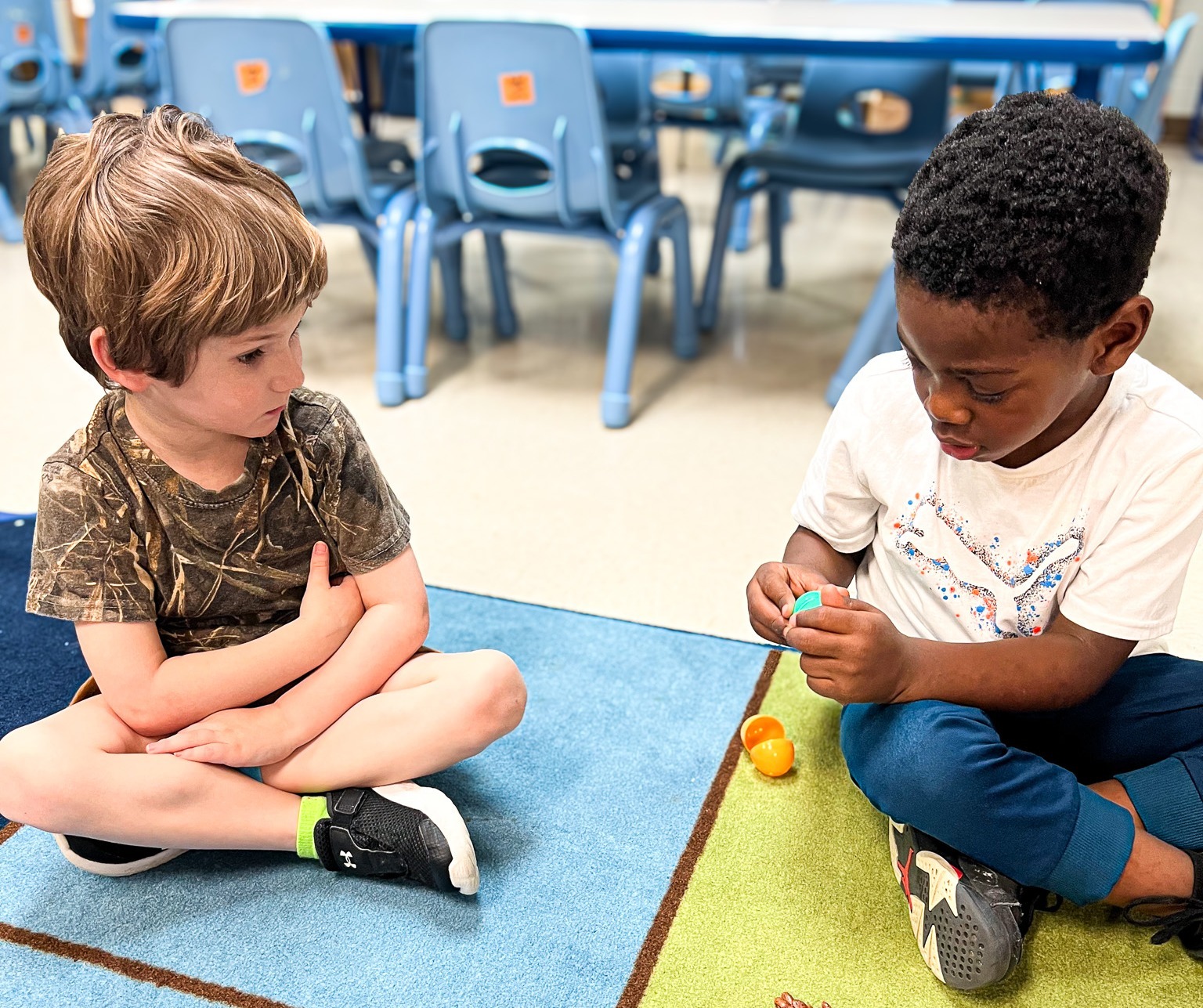 Two children sit on the floor, one with a bracelet, sorting objects on a colorful rug. Behind them, blue chairs and a table are visible.