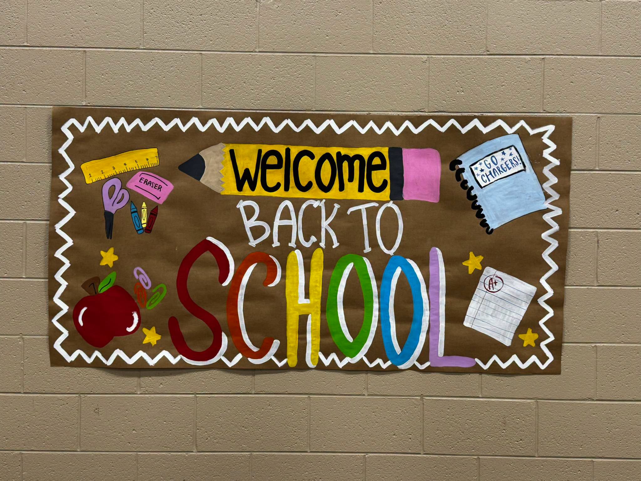 Decorative banner on a brick wall. It reads "Welcome Back to School" in colorful letters. Items like a pencil, scissors, and books adorn the banner.