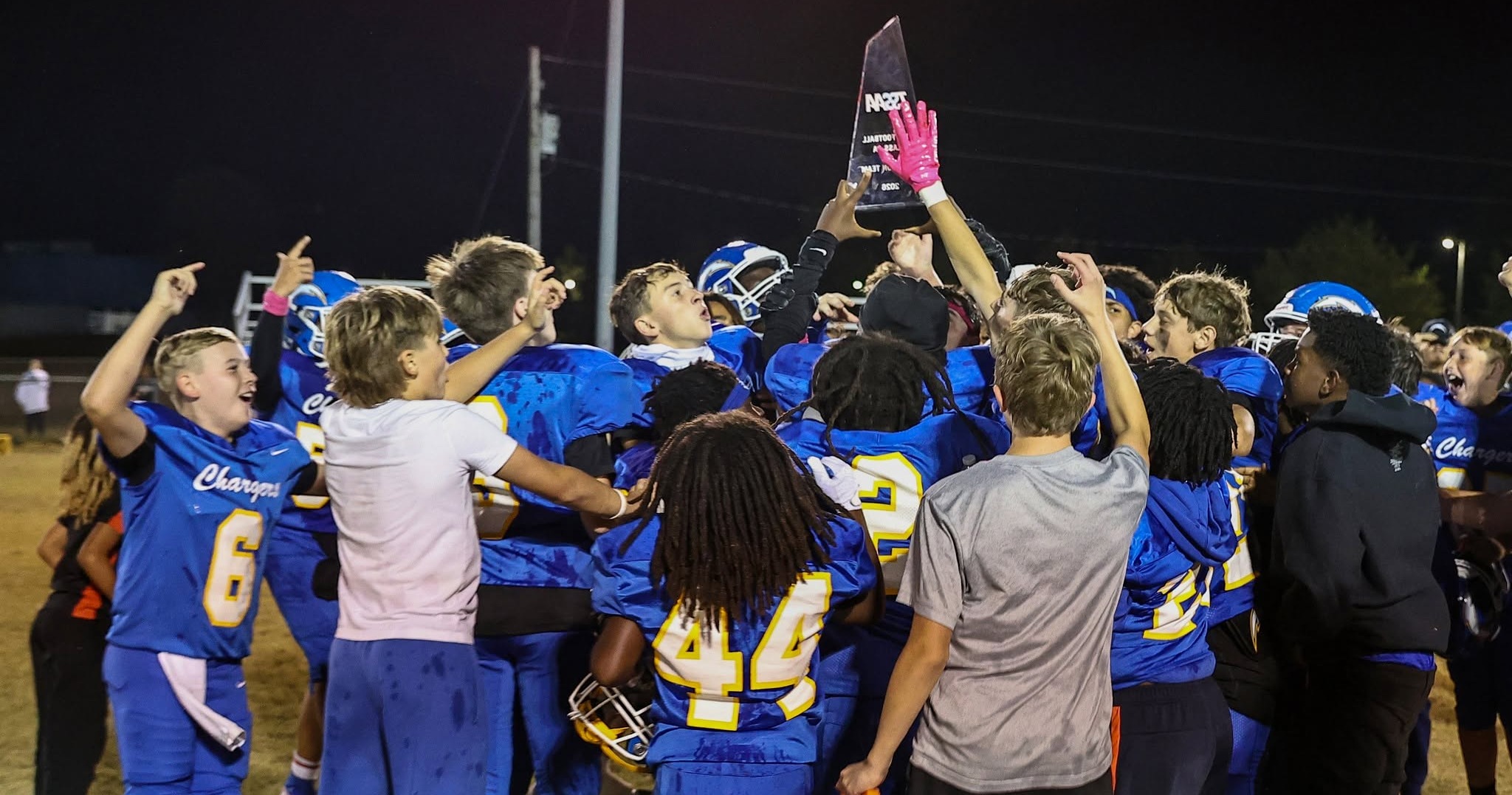 A group of football players in blue uniforms celebrate with a trophy and a bag on a field.