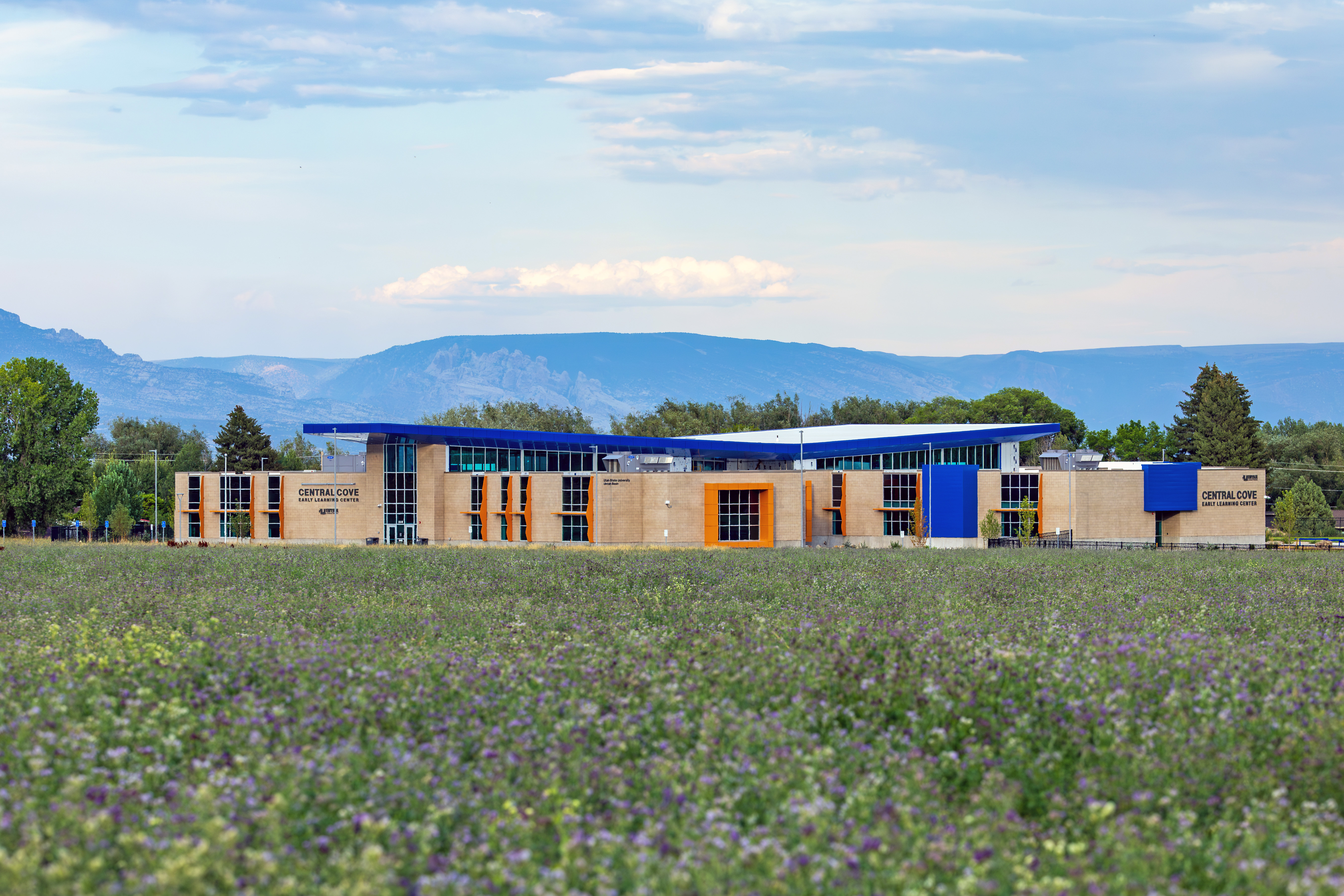 A modern, tan-and-blue building labeled “Central Cove Early Learning Center” sits beyond a wide field of alfalfa, with green trees in the background and distant blue mountains under a partly cloudy sky.