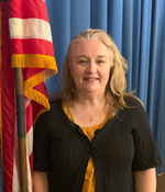A woman with long, wavy hair stands in front of a blue curtain and an American flag, wearing a black cardigan over a yellow top.
