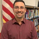 A smiling man wearing glasses and a maroon shirt stands in front of a bookshelf, with an American flag in the background.