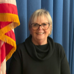 A woman with short blonde hair and glasses smiles while standing next to an American flag, against a blue curtain backdrop.