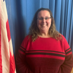 A woman with long, curly hair wearing a red sweater with black stripes stands in front of a blue curtain and an American flag.