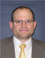 A man with glasses and a light brown beard, wearing a gray suit and a yellow tie, poses for a professional headshot against a blue background.