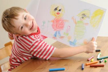 A smiling child in a striped shirt proudly holds up a colorful drawing featuring two cartoonish figures, surrounded by crayons on a wooden table.