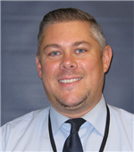 A smiling man with short hair and a goatee, wearing a light blue dress shirt and a dark tie, poses against a dark background.