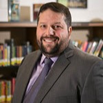 Man with beard & ustache in a grey suit in front of a bookcase