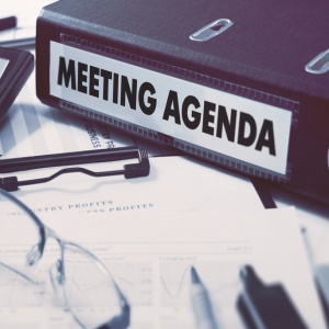 A close-up view of a binder labeled 'MEETING AGENDA' placed on a desk, surrounded by papers, a clipboard, and a pair of glasses.