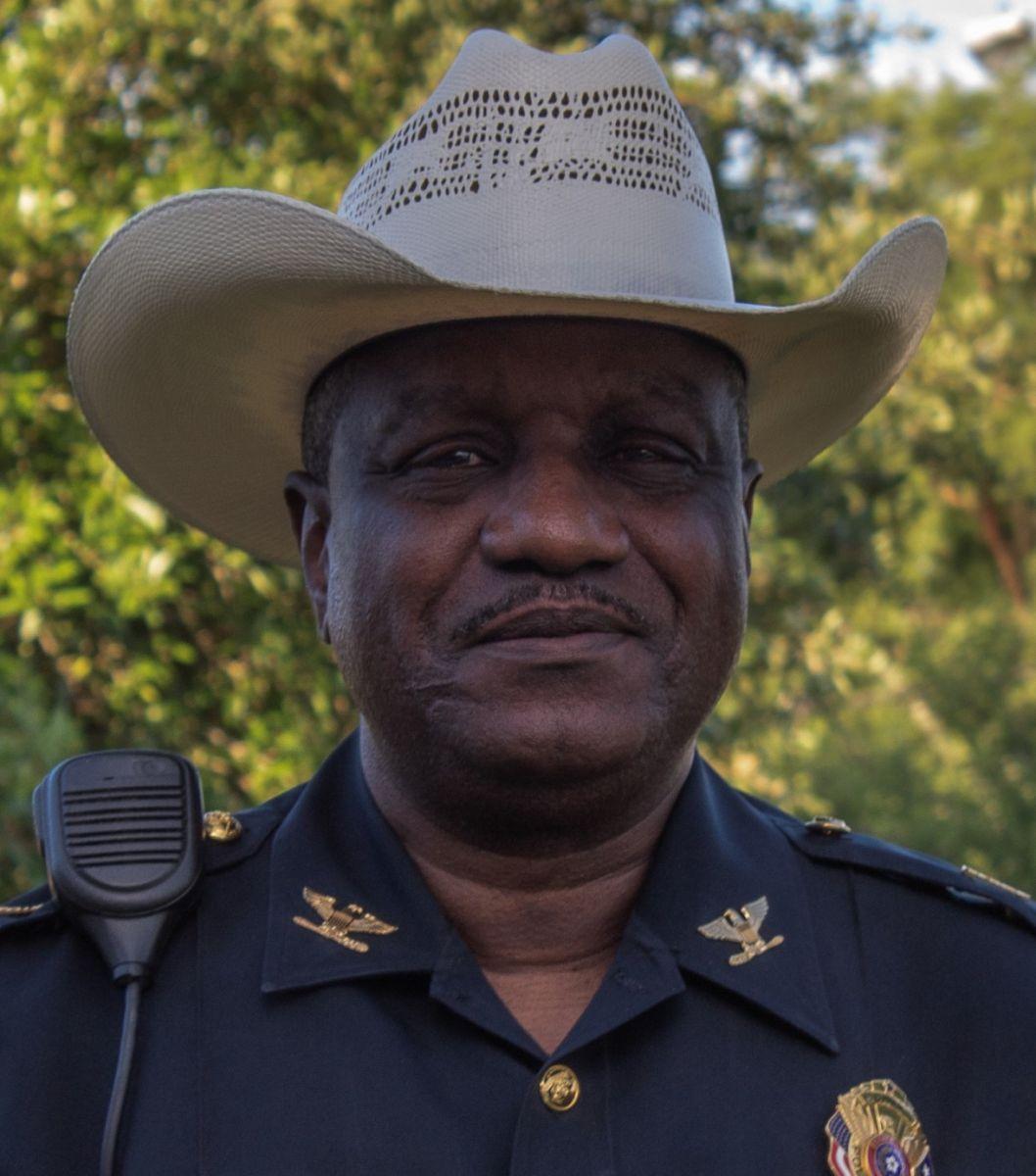 A man in a police uniform with a cowboy hat stands outdoors, smiling at the camera.