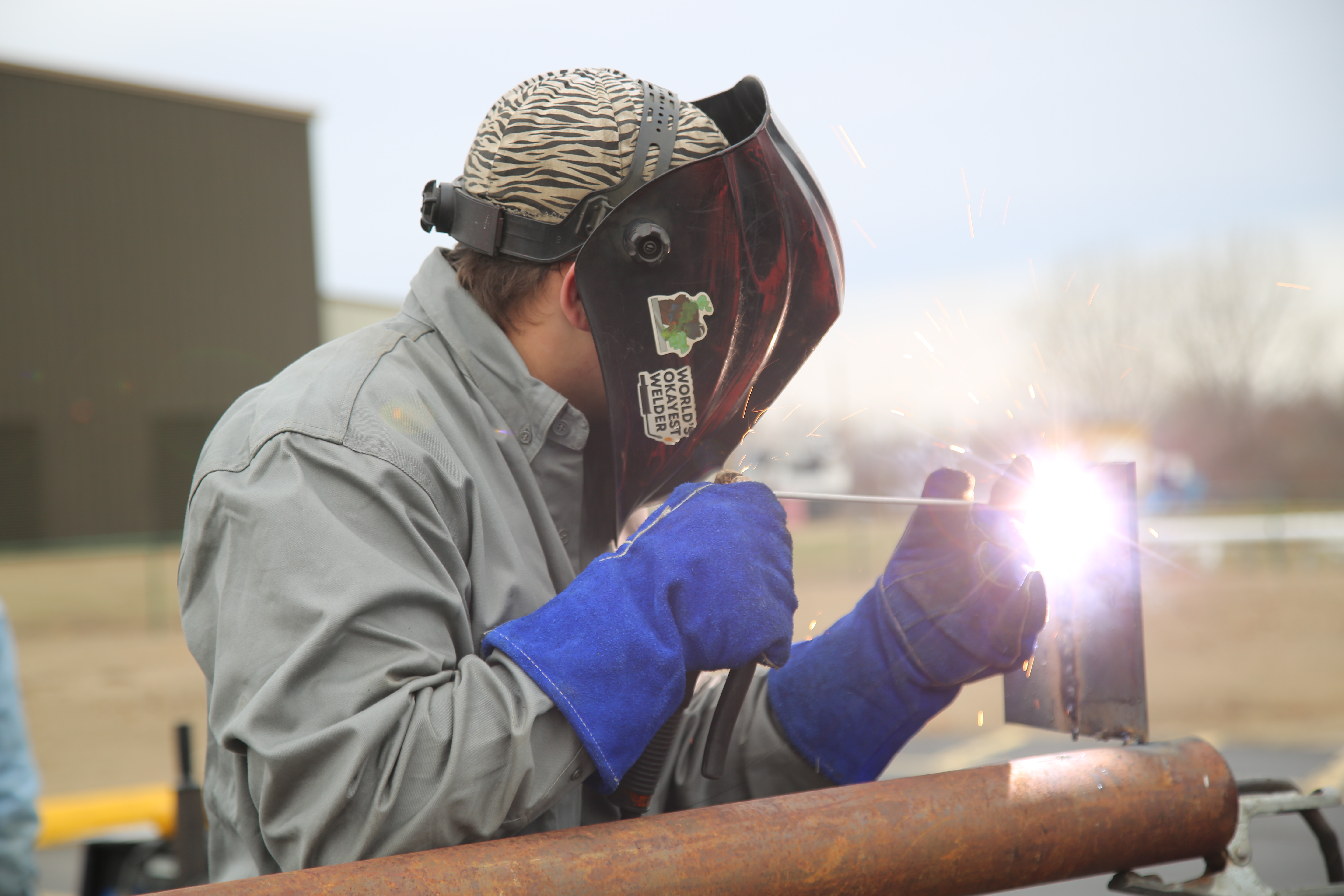 A welder wearing protective gear and a helmet is focused on welding a metal piece, with a bright spark visible at the welding point.