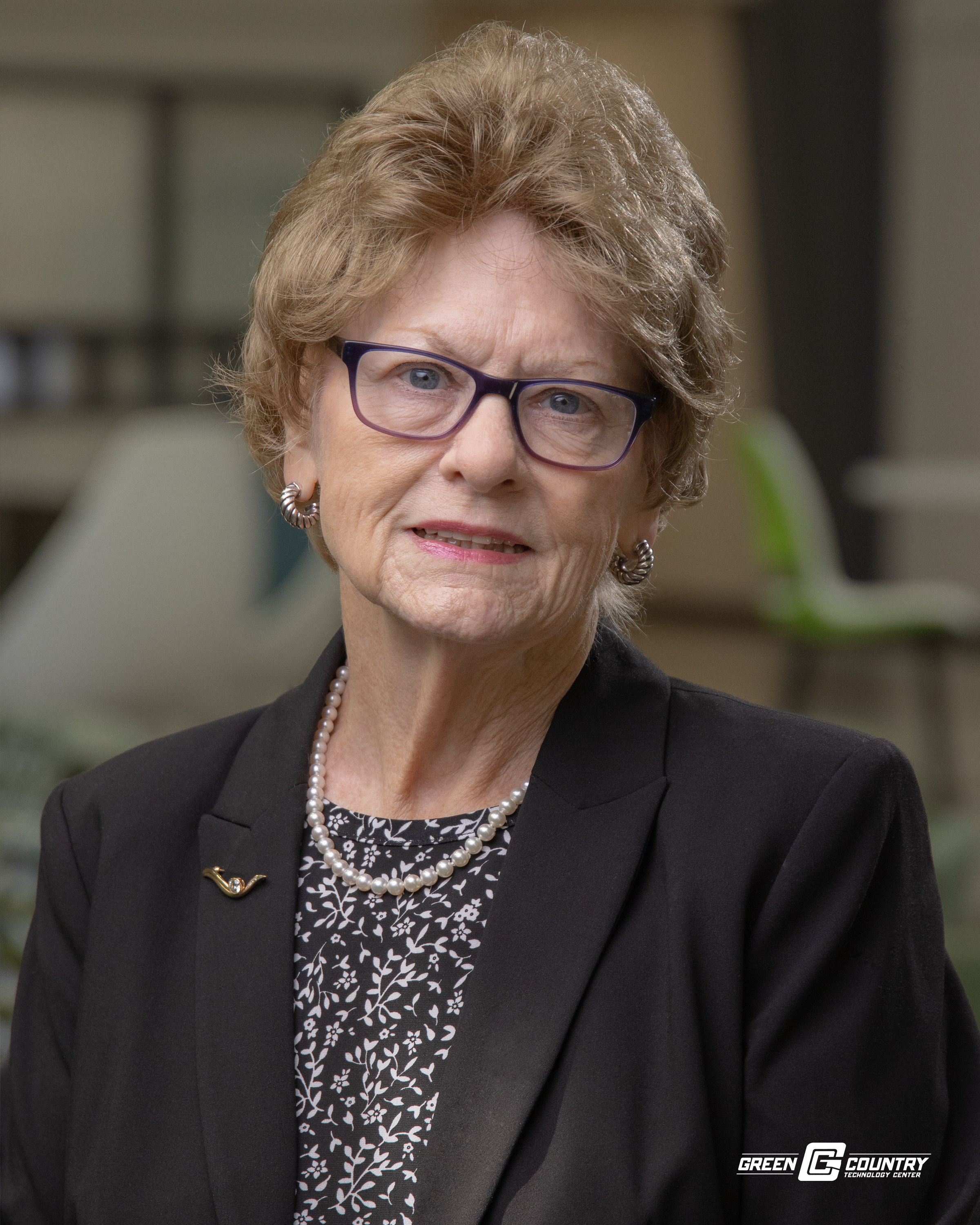 A woman with short, curly hair and glasses is wearing a black blazer over a patterned blouse, posing for a portrait in a well-lit indoor setting with modern furniture in the background.
