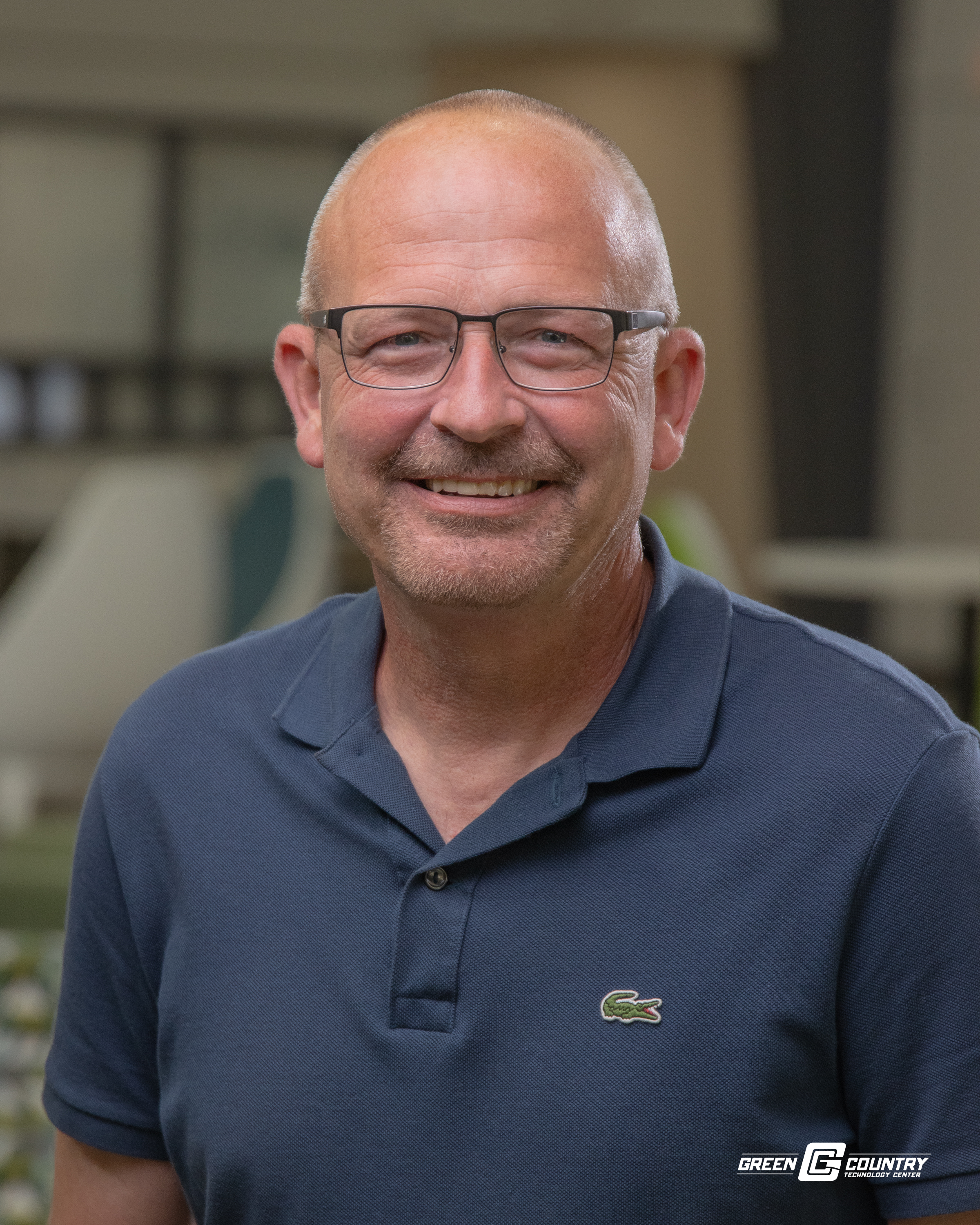 A smiling man with glasses and a short beard, wearing a navy blue polo shirt, stands in a well-lit indoor setting with blurred background elements.