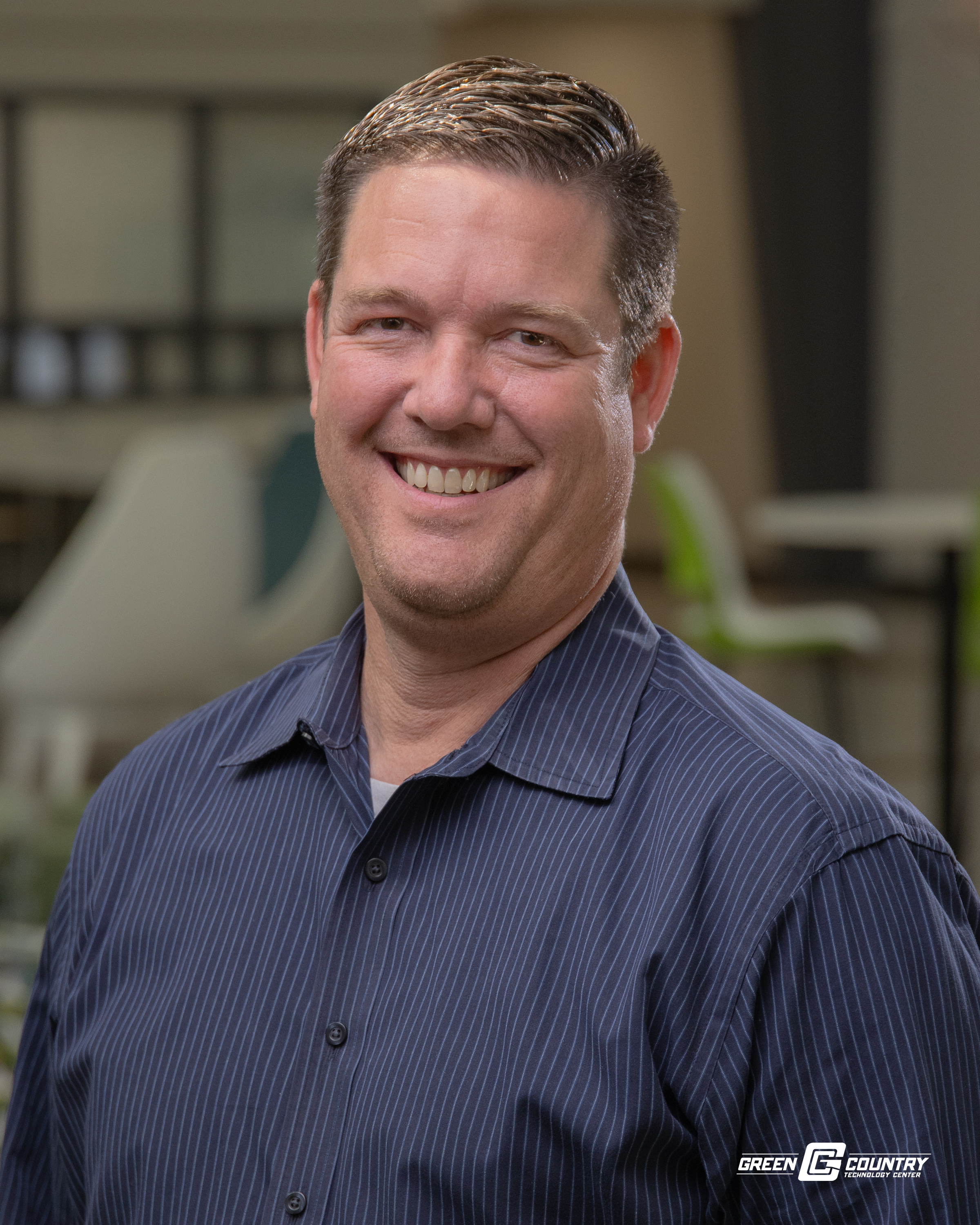 A smiling man with short hair, wearing a dark blue striped shirt, stands in an indoor setting with soft lighting and blurred background elements.