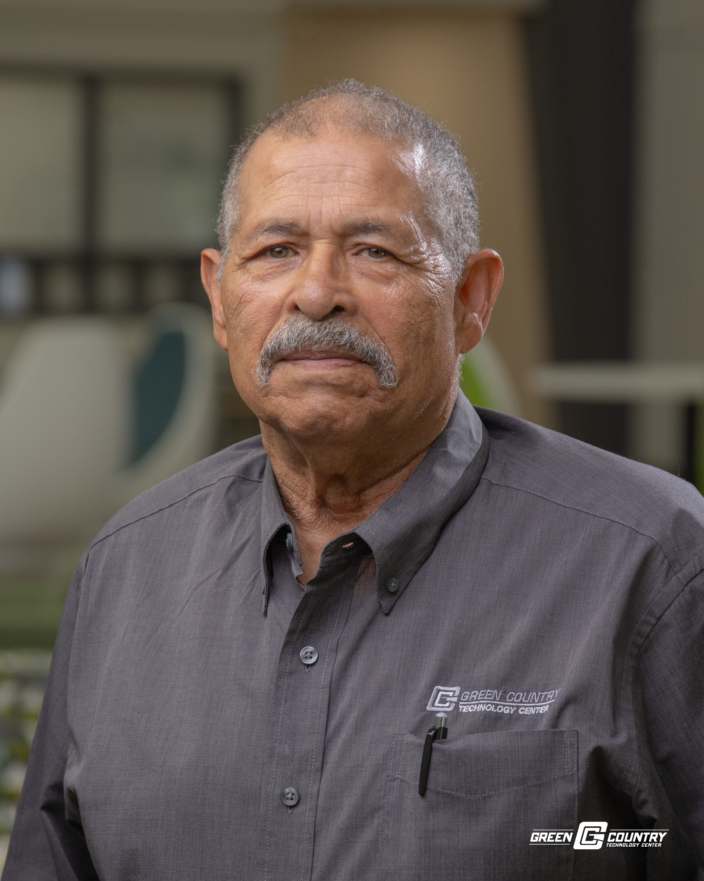A senior man with a mustache and short gray hair, wearing a gray button-up shirt, stands in front of a blurred background, likely indoors.
