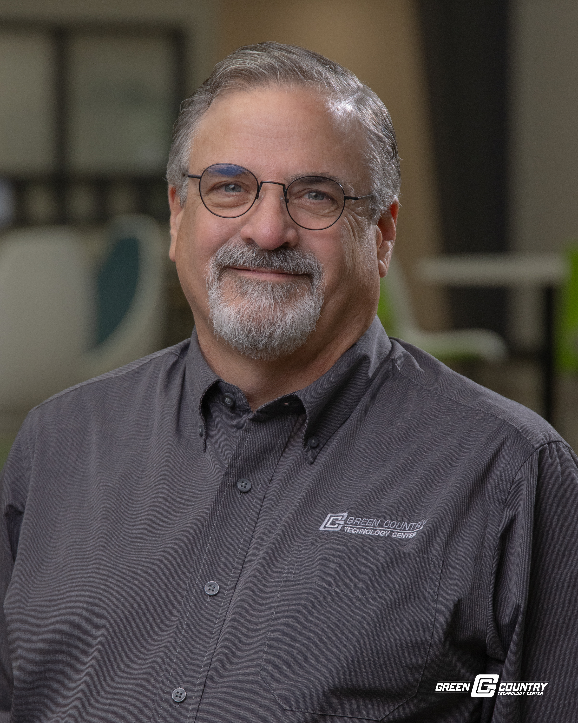 A middle-aged man with glasses and a beard smiles while wearing a gray collared shirt with a logo on the left side.