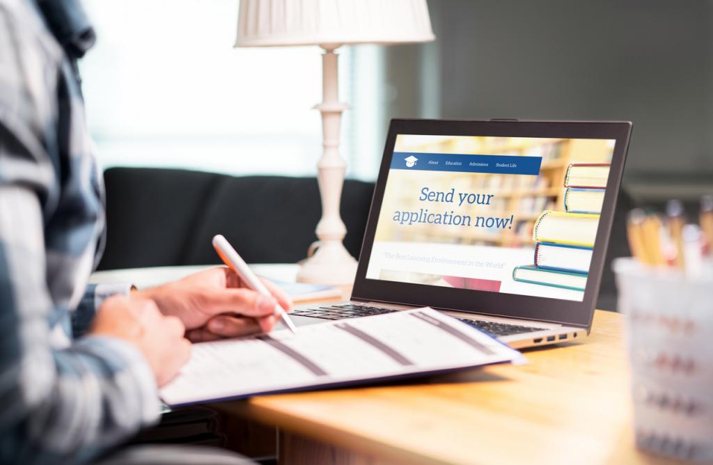 A person is sitting at a desk, writing on a notepad while looking at a laptop screen that displays a message encouraging users to send their application.