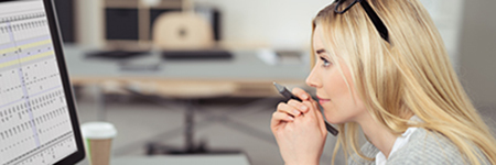 A woman with long blonde hair is sitting at a desk, thoughtfully looking at a computer screen while holding a pen to her lips.