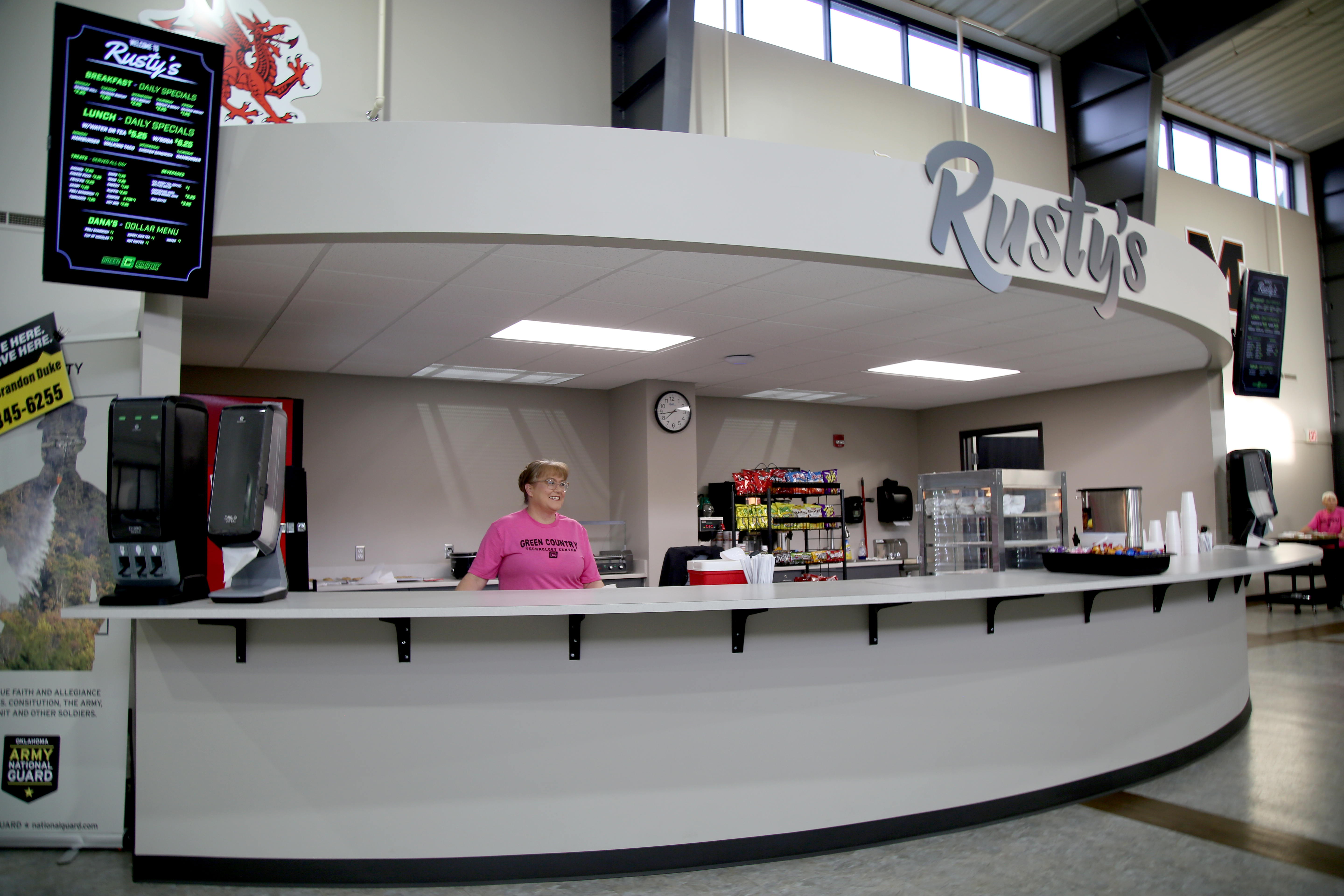 A concession stand named 'Rusty's' featuring a counter with a staff member in a pink shirt, various food and drink items displayed, and digital menu boards on the walls.