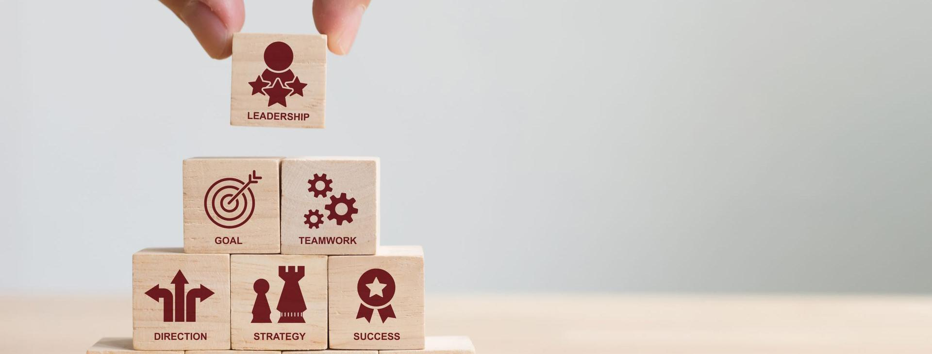 Hand placing a wooden block labeled 'Leadership' on top of a pyramid made of other blocks representing key concepts like communication, direction, strategy, teamwork, and vision—symbolizing the building blocks of effective leadership.