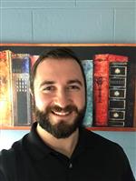A smiling man with a beard poses in front of a colorful bookshelf painting.