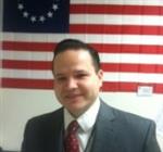 A man in a suit stands in front of a large American flag with 13 stars and red and white stripes.