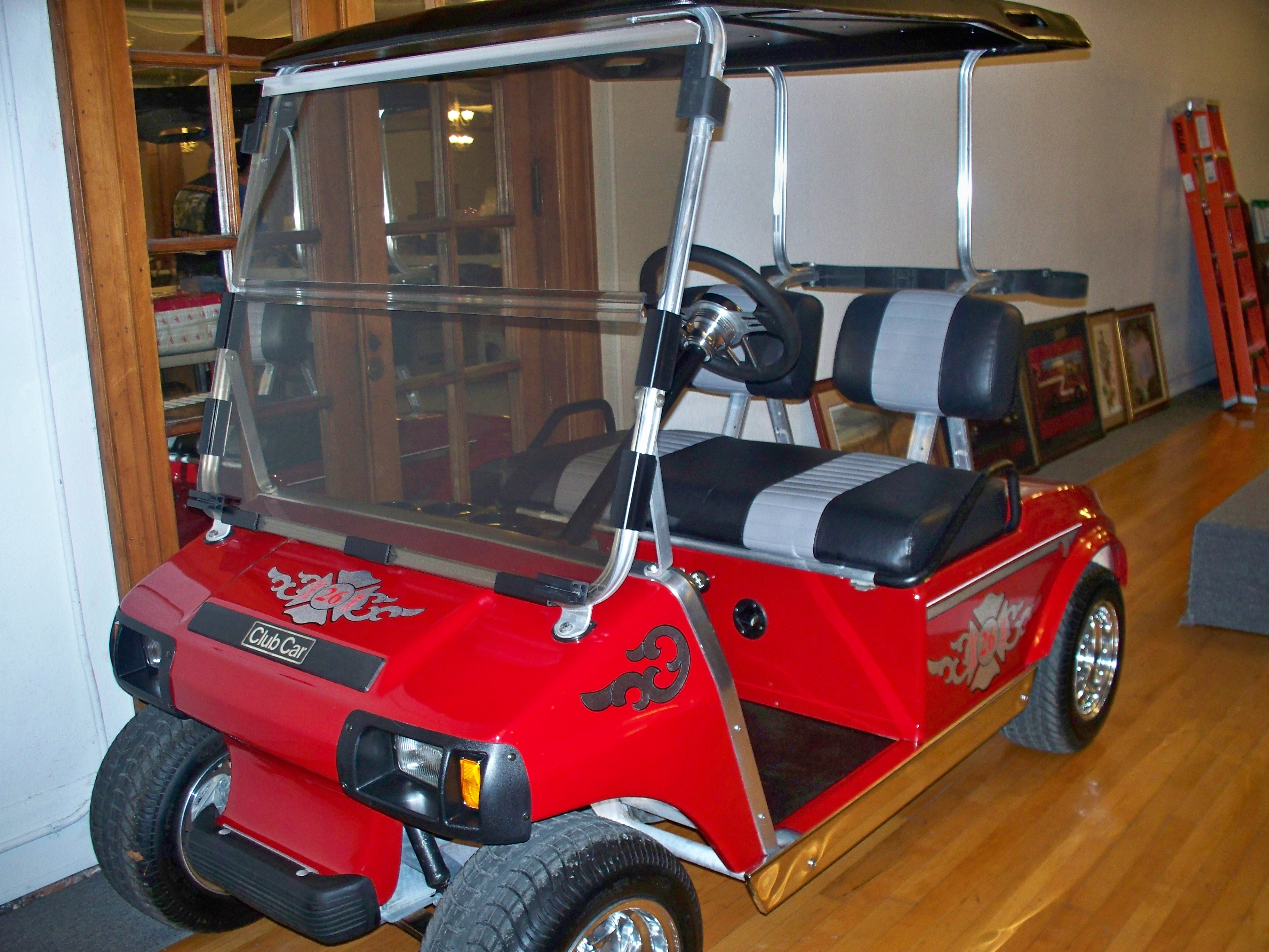 A red golf cart with a black roof and gray seats, parked indoors with wooden flooring and shelves in the background.