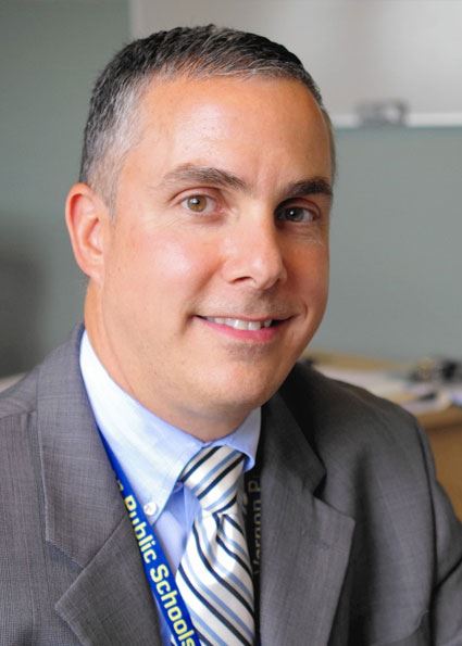 A man in a suit and tie smiles at the camera, with a school environment in the background.