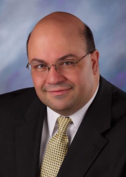 A man with a bald head and glasses, wearing a black suit and a light-colored tie, smiles at the camera against a soft, blurred background.