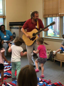 A man playing guitar engages with children dancing in a lively indoor setting, with adults observing in the background.