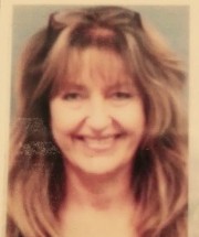 A smiling woman with long, wavy hair and bangs, wearing a light-colored top, against a soft background.