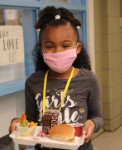 A young girl wearing a pink mask and a gray shirt with the text 'Girls Rule' smiles while holding a tray of food, which includes a sandwich, a drink, and some vegetables, in a school setting.