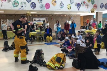 A firefighter in uniform stands in front of a group of children seated on the floor, while others are gathered around tables decorated for an event, with colorful artwork hanging on the walls.