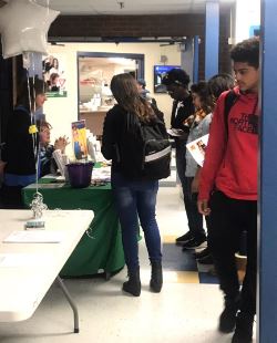 A group of students gathers around a table in a school hallway, engaging with a representative while others wait in line, with informational materials displayed on the table.