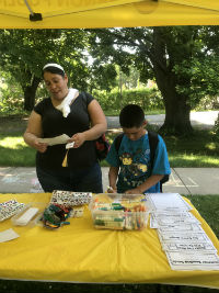 A woman and a boy are standing at a table under a yellow canopy, with various items displayed on the table, including snacks and papers, in a park-like setting.