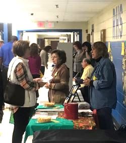 A group of people engaging in conversation at tables set up in a school hallway during an event, with various displays and attendees in the background.