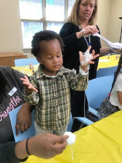 A young child with curly hair is experimenting with a sticky substance, looking focused while holding it in both hands, surrounded by adults in a brightly lit room with yellow tables.