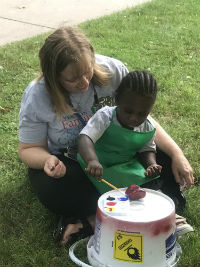 A woman and a child are sitting on the grass, with the child painting on a white bucket using a paintbrush, while the woman observes and assists.