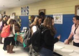 A busy school hallway with students interacting at tables, some carrying backpacks, while a staff member assists at a booth.