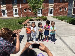 A group of four children stands together outdoors, holding colorful items, while an adult takes their photo with a smartphone.