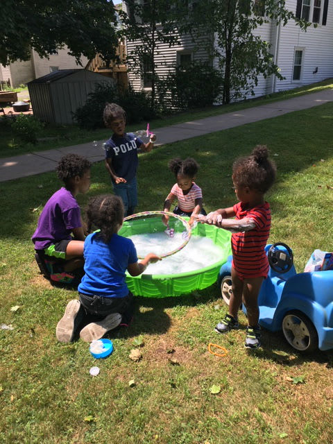 A group of children playing with a large, green inflatable pool in a grassy yard on a sunny day, with one child holding a bubble wand and others engaged in the activity.