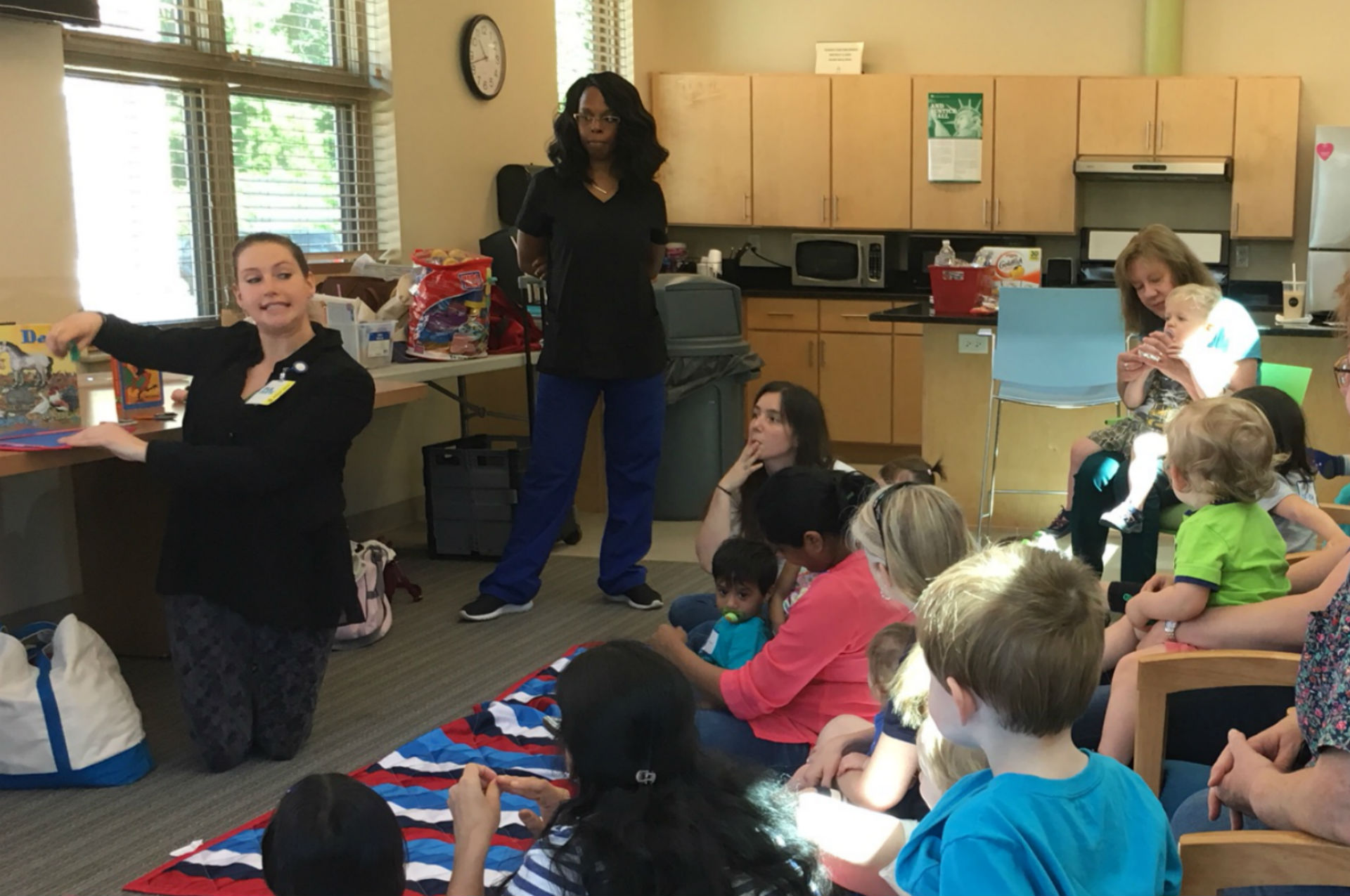 A group of children and adults are gathered in a room, with one woman sitting on the floor demonstrating an activity while another woman stands nearby, all engaged in a learning session.