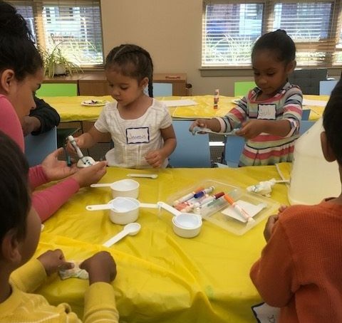 A group of young children engaged in a creative activity at a table covered with a yellow cloth, using various art supplies and materials.
