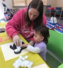 A woman and a young child are engaged in a craft activity, with the woman helping the child place cotton balls on a black cutout on a table covered with yellow paper.