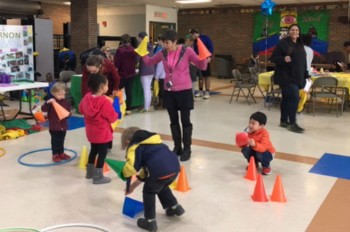 A group of children play with colorful cones and hula hoops in a community center, while adults supervise and engage in the background.