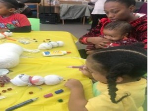 A woman holds a baby while a child in a yellow shirt paints a snowman made of white balls on a yellow table, surrounded by craft materials.