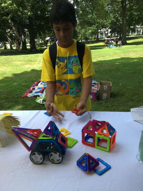 A child in a yellow shirt is playing with colorful magnetic building blocks on a table in a park, with a toy vehicle and a house structure made from the blocks in front of him.
