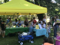 A lively outdoor event featuring a yellow tent with people gathered around various tables, engaging in activities and enjoying the day.